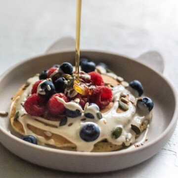 A stack of pancakes, covered in fruit, with maple syrup being poured over.
