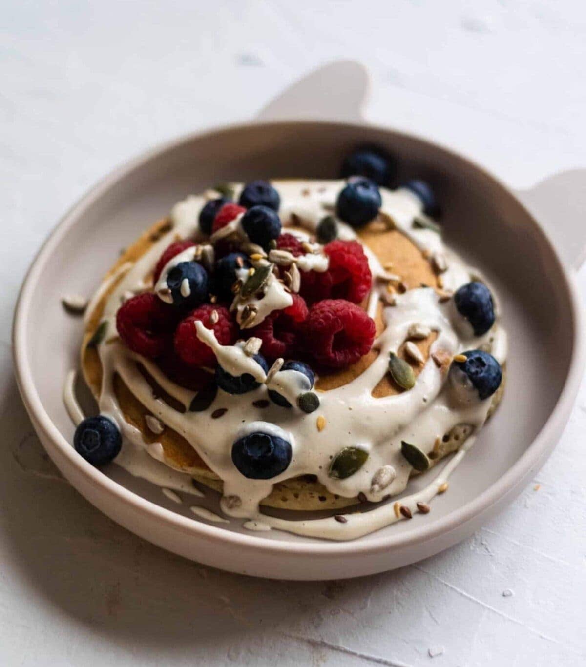 A plate of pancakes topped with cashew cream, blueberries, raspberries, sunflower and pumpkin seeds.