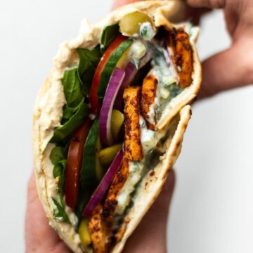 Someone holding a pitta, filled with Greek tofu and salad, drizzled with tzatziki. Holding it in both hands, the camera looking down on it.