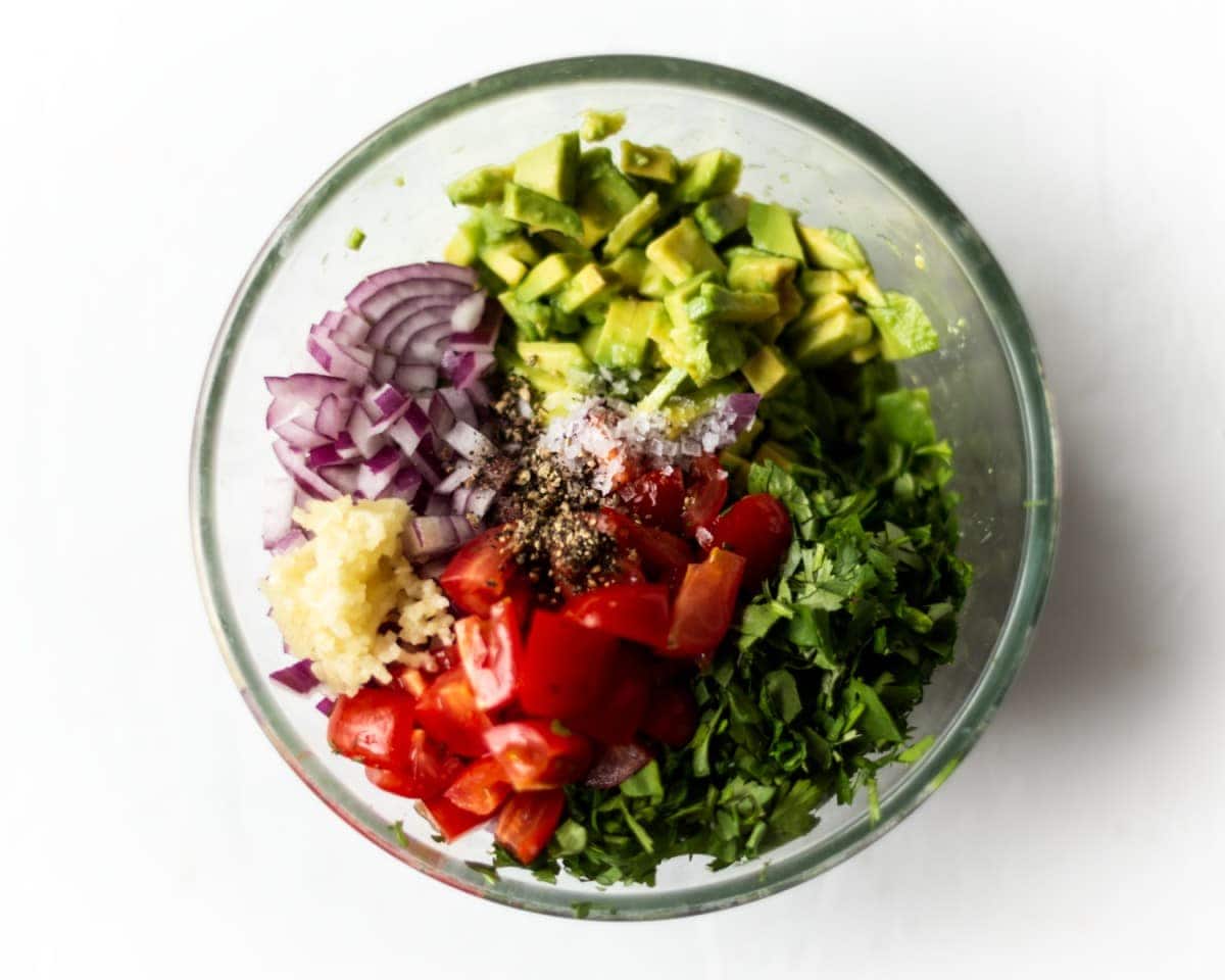 All for he guacamole ingredients together in a glass bowl, prior to being mixed together. photographed from above.