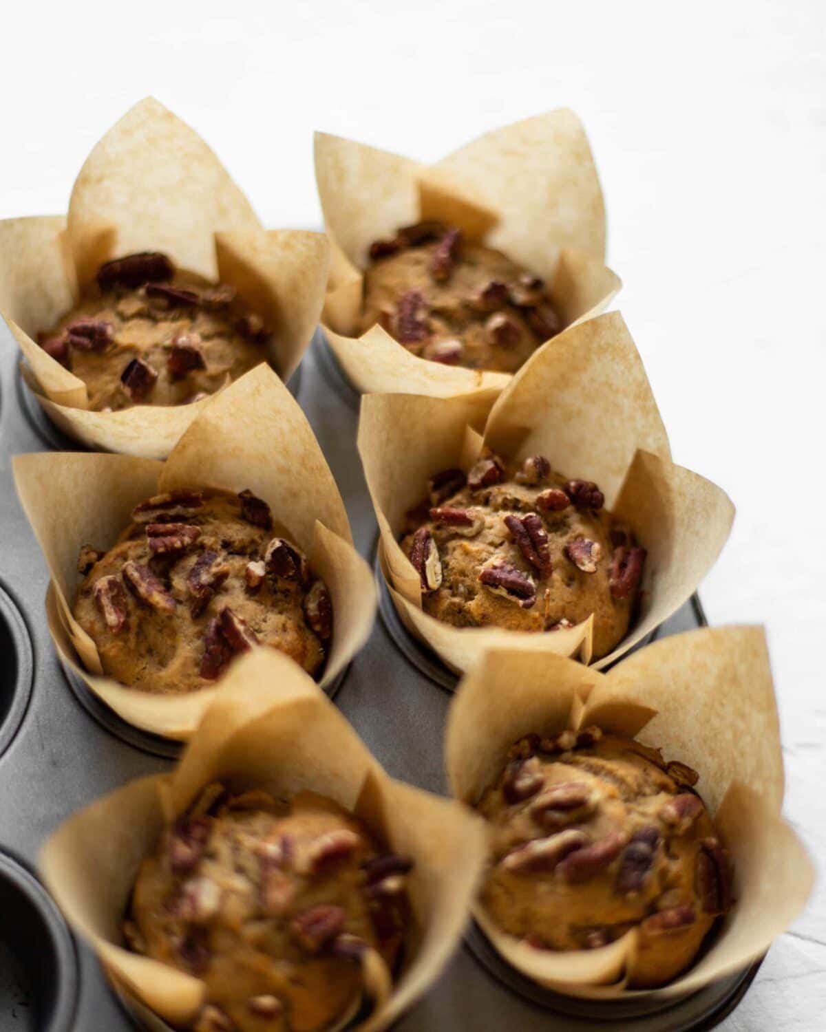 A muffin tray full of baked muffins, in large brown paper muffin cases, topped with pecans.