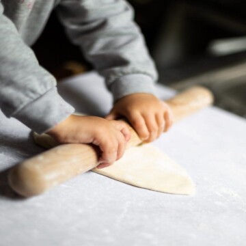 Toddler rolling out the pasta dough on a work surface, using a rolling pin.