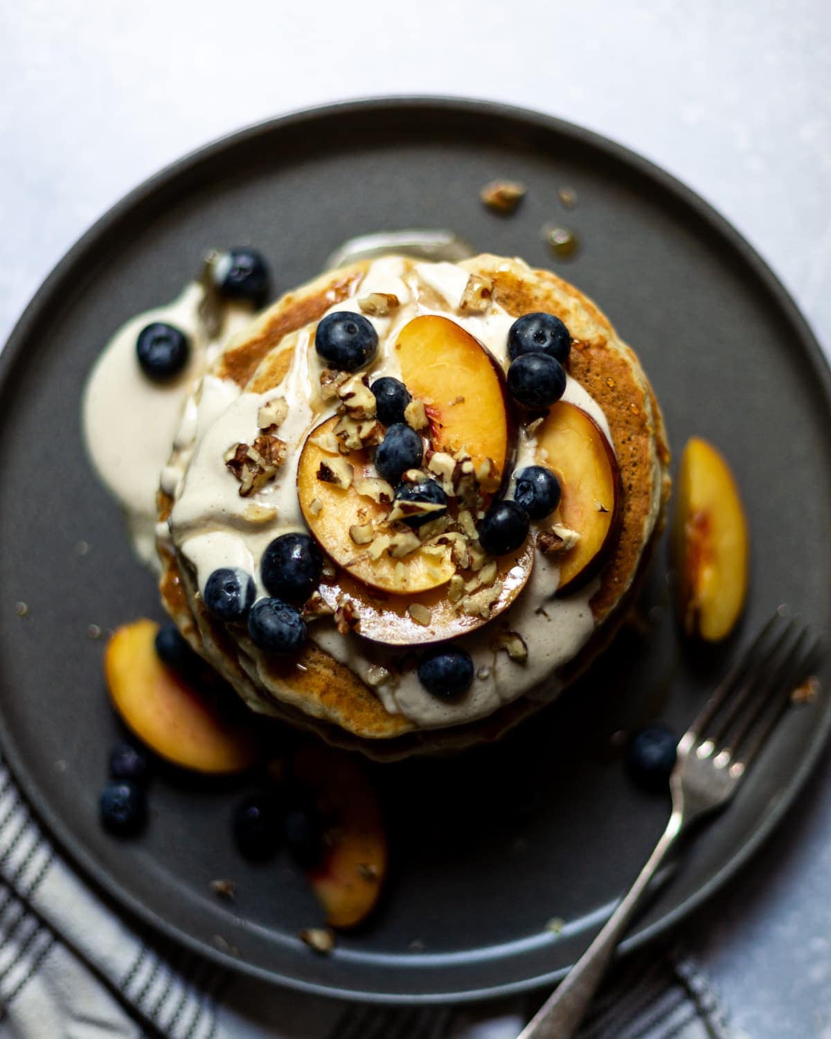 Overhead view of a stack of fluffy pancakes topped with cashew cream, sliced peaches, blueberries, chopped walnuts and maple syrup.