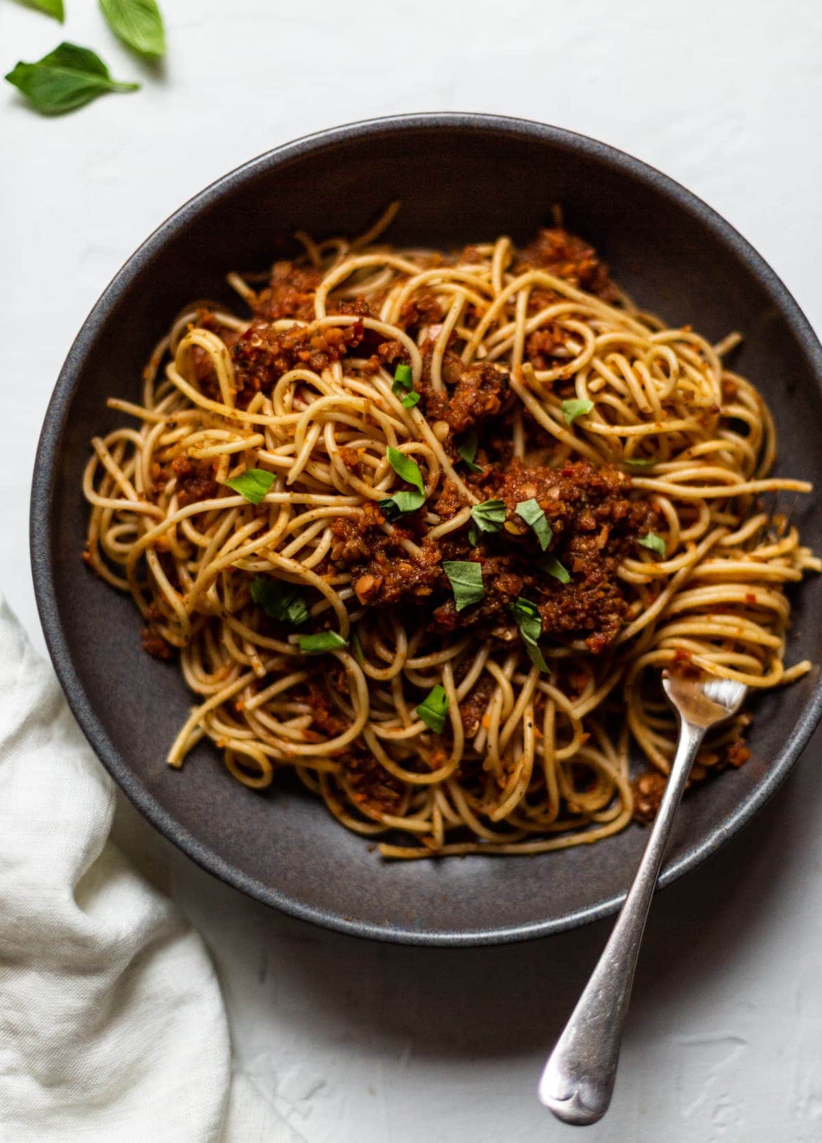 A grey bowl on a white background filled with our mushroom and lentil bolognese and spaghetti pasta. Topped with chopped fresh basil.