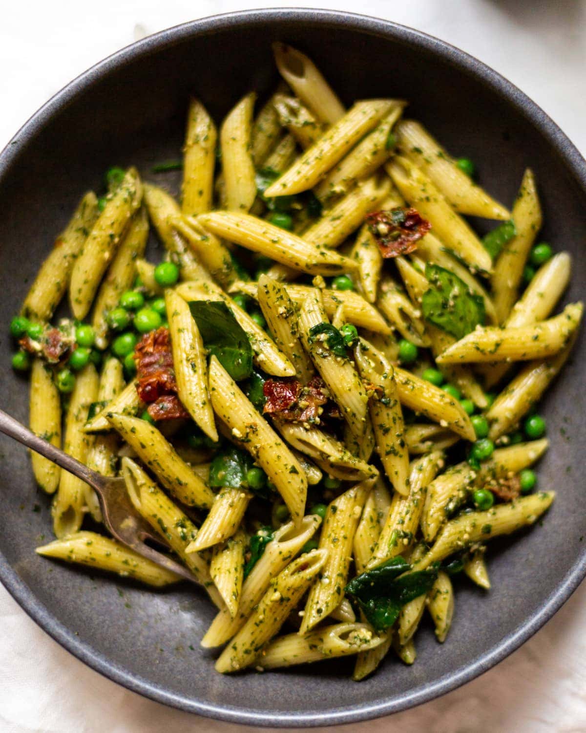 An overhead photo of penne pesto pasta in a dark grey bowl, on a white linen tablecloth.