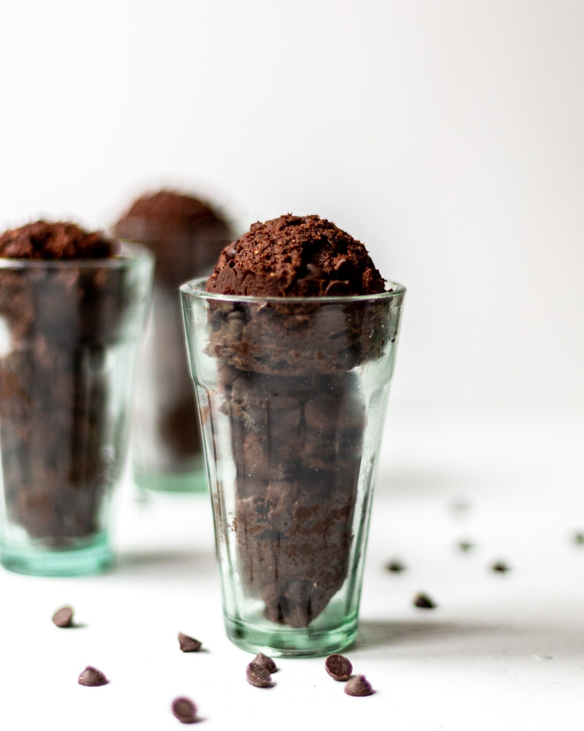 Three scoops of double chocolate, edible cookie dough, in a tall glass, made to look like ice-cream, on a white background with scattered chocolate chips in the foreground.