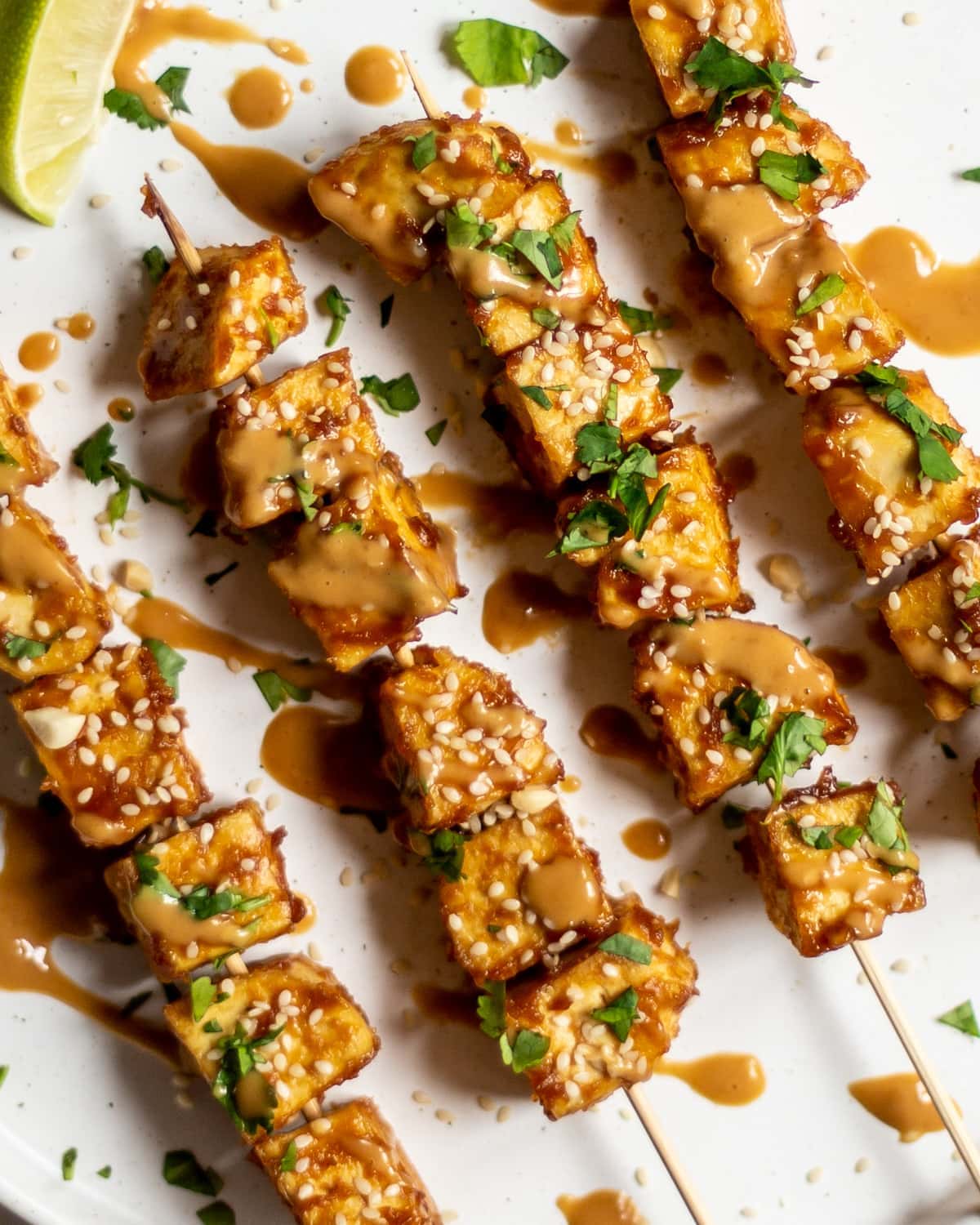 A close up of four satay tofu skewers on a white plate, on a white background, garnished with fresh coriander and sesame seeds, and drizzled with peanut sauce.