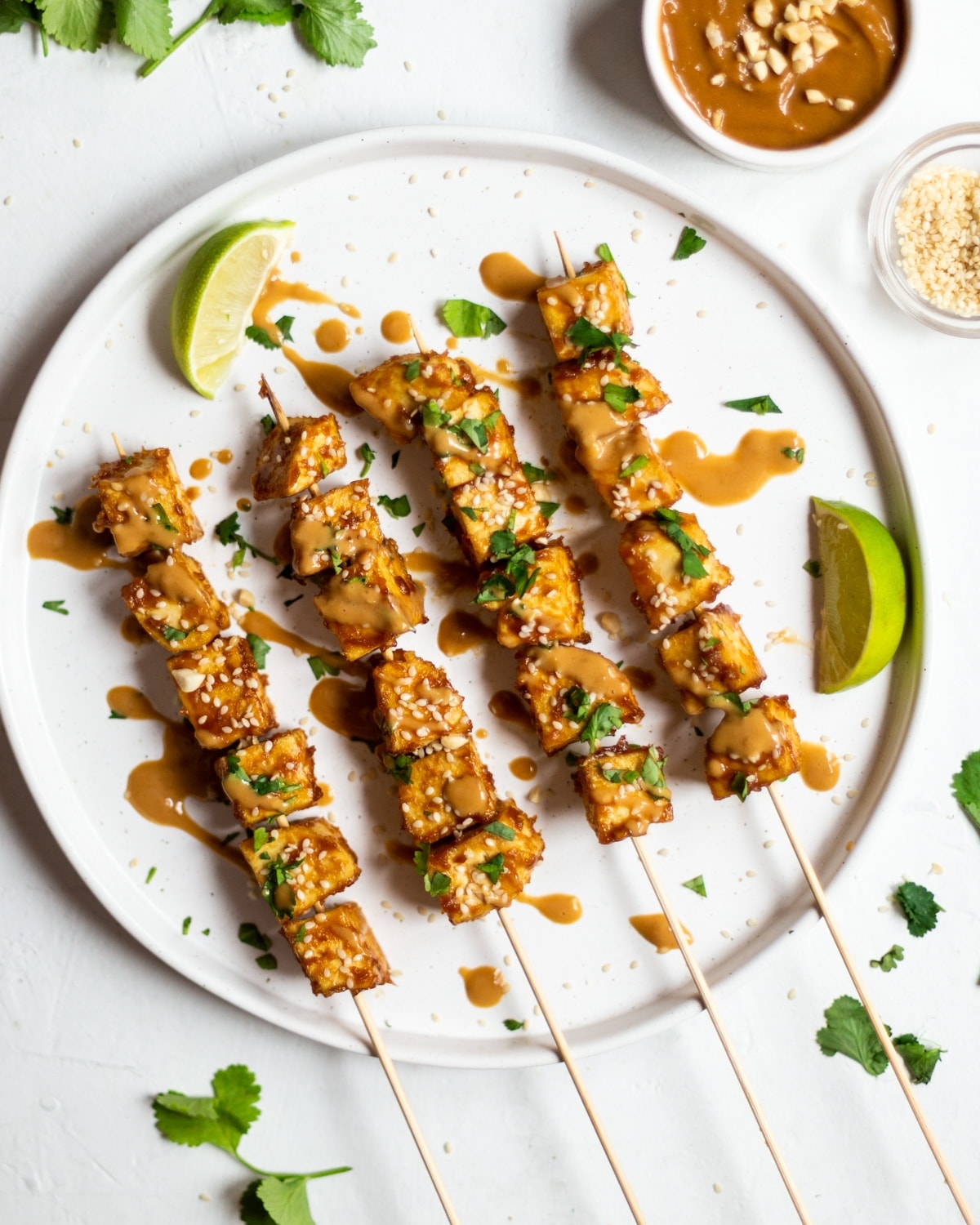 Four satay tofu skewers on a white plate, on a white background. Garnished with fresh coriander and sesame seeds, drizzled with peanut sauce.