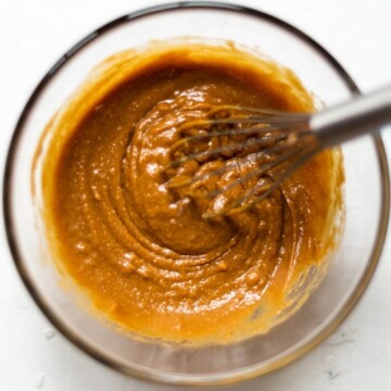 A glass bowl full of Thai style vegan satay sauce, on a white background, with a whisk in the centre of the bowl.