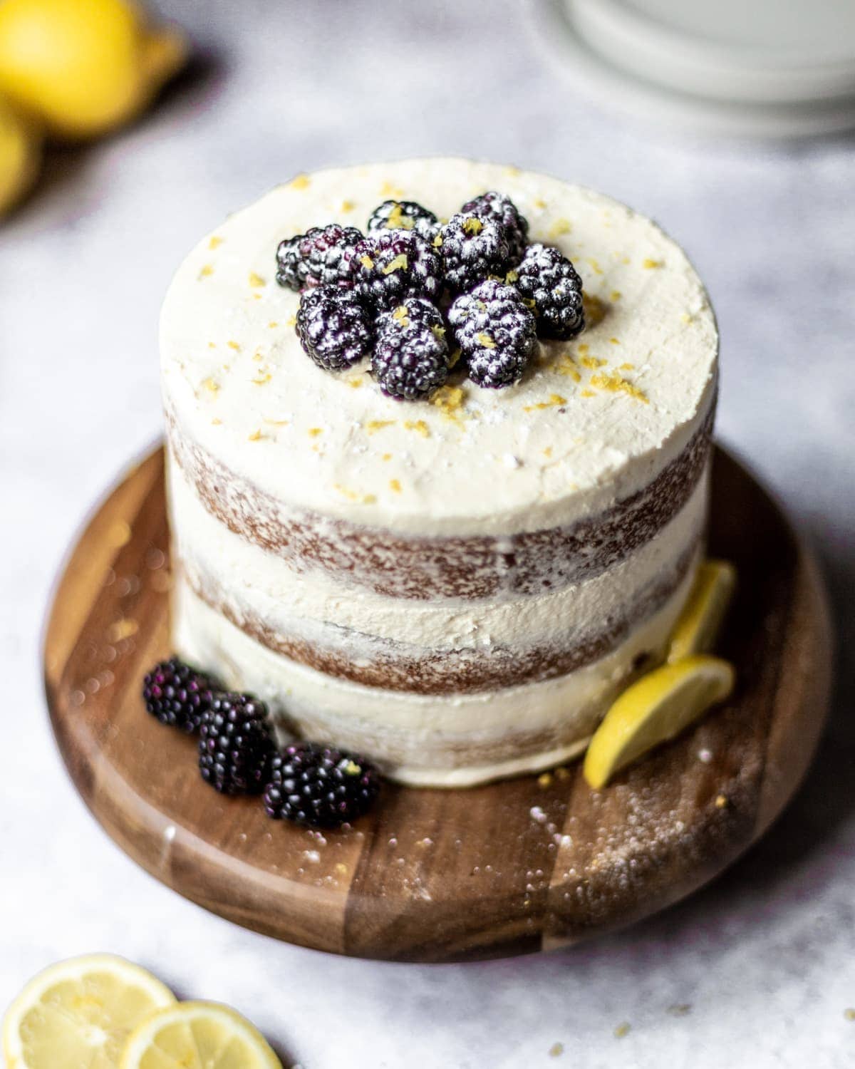 A whole triple layered lemon blackberry cake, stood on a wooden board, decorated with buttercream, blackberries and lemon zest and slices.
