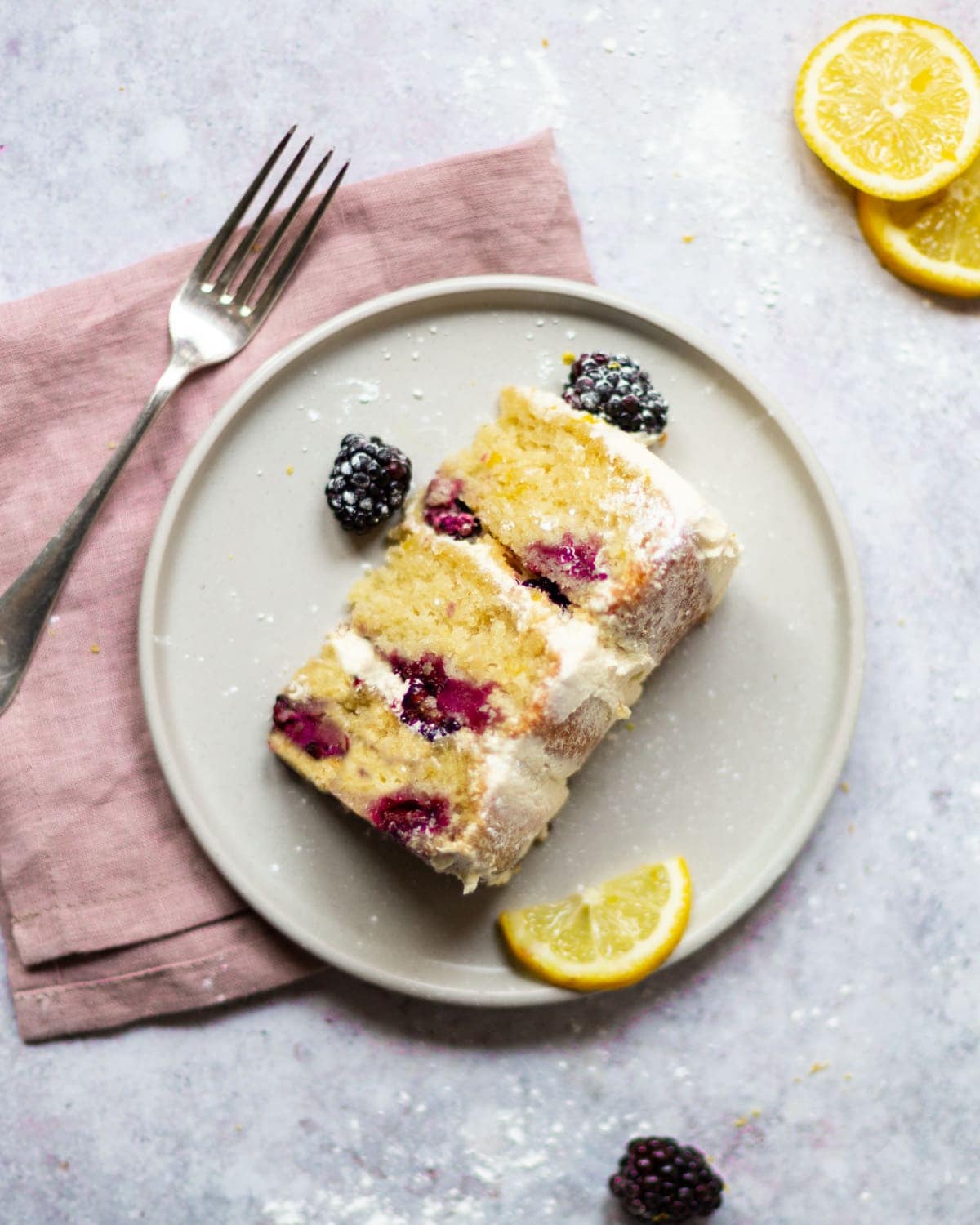 An over head shot of a slice of lemon blackberry cake on a plate. The plate is sat on a pink napkin, with a fork sat next to it.