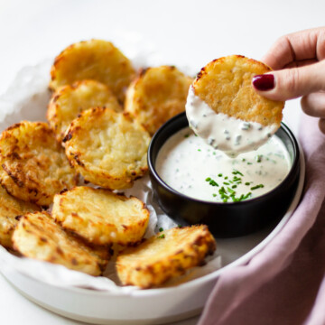 Side angle of a white bowl filled with the baked hash browns, and a black bowl filled with chive dip. One has brown is being dipped 1/3 of the way in to the chive dip.