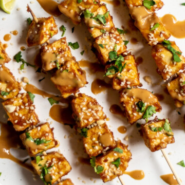 A close up of four satay tofu skewers on a white plate, on a white background, garnished with fresh coriander and sesame seeds, and drizzled with peanut sauce.